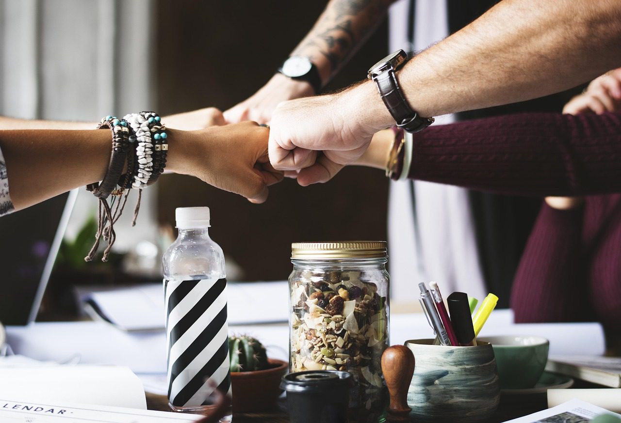 Hands of diverse individuals fist-bumping, symbolizing teamwork and collaboration at a meeting table with snacks, stationery, and a water bottle.
